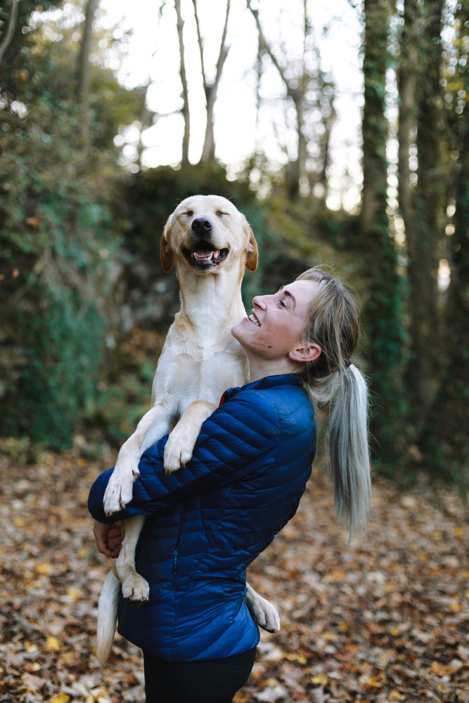 Large cream-colored dog being picked up by woman in the woods, both smiling
